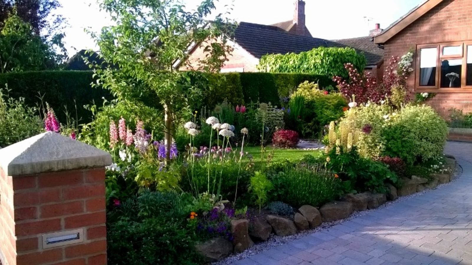 Front garden in full bloom with lupins, alliums and stone-edged borders beside a block-paved drive