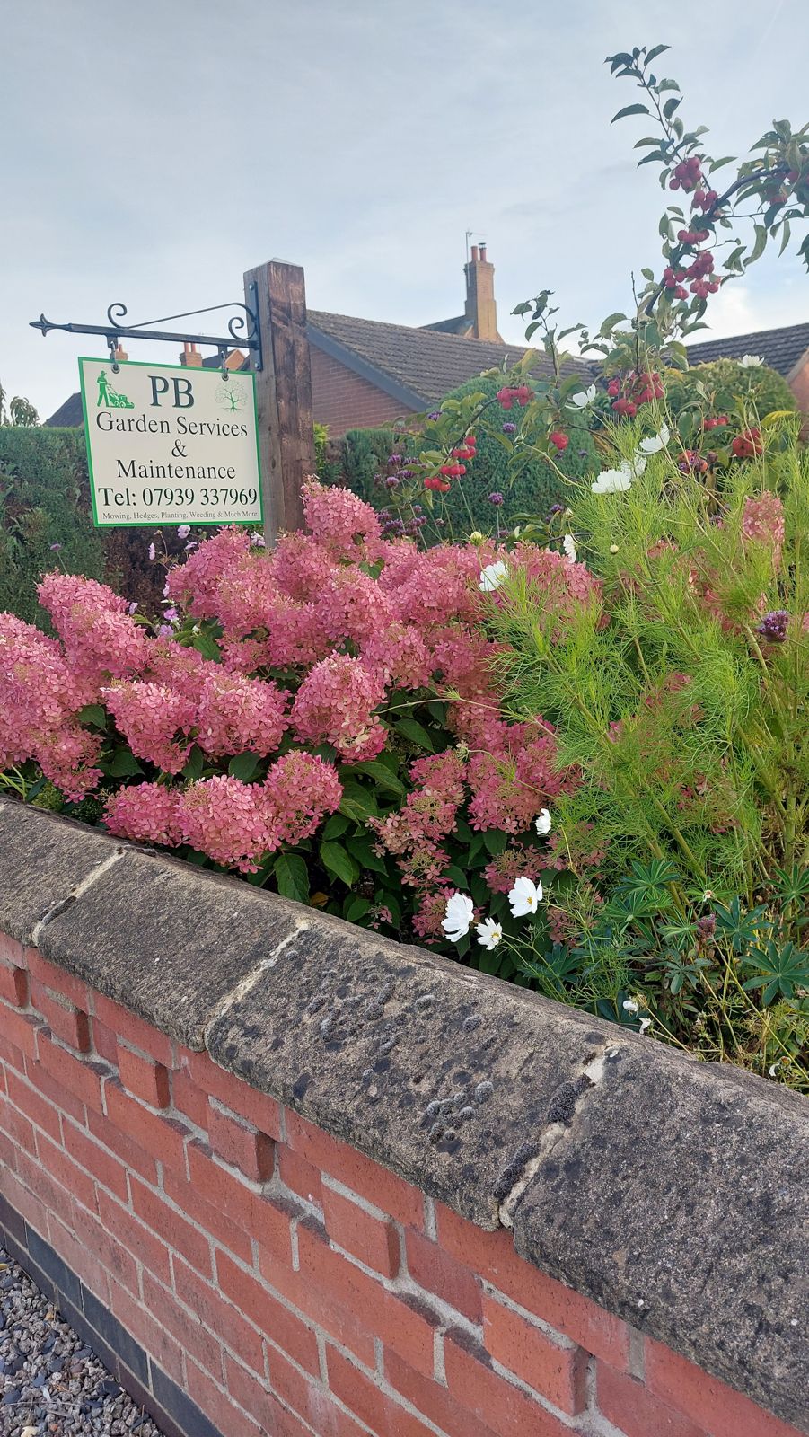 The PB Garden Services & Maintenance hanging sign above pink hydrangeas and a brick wall